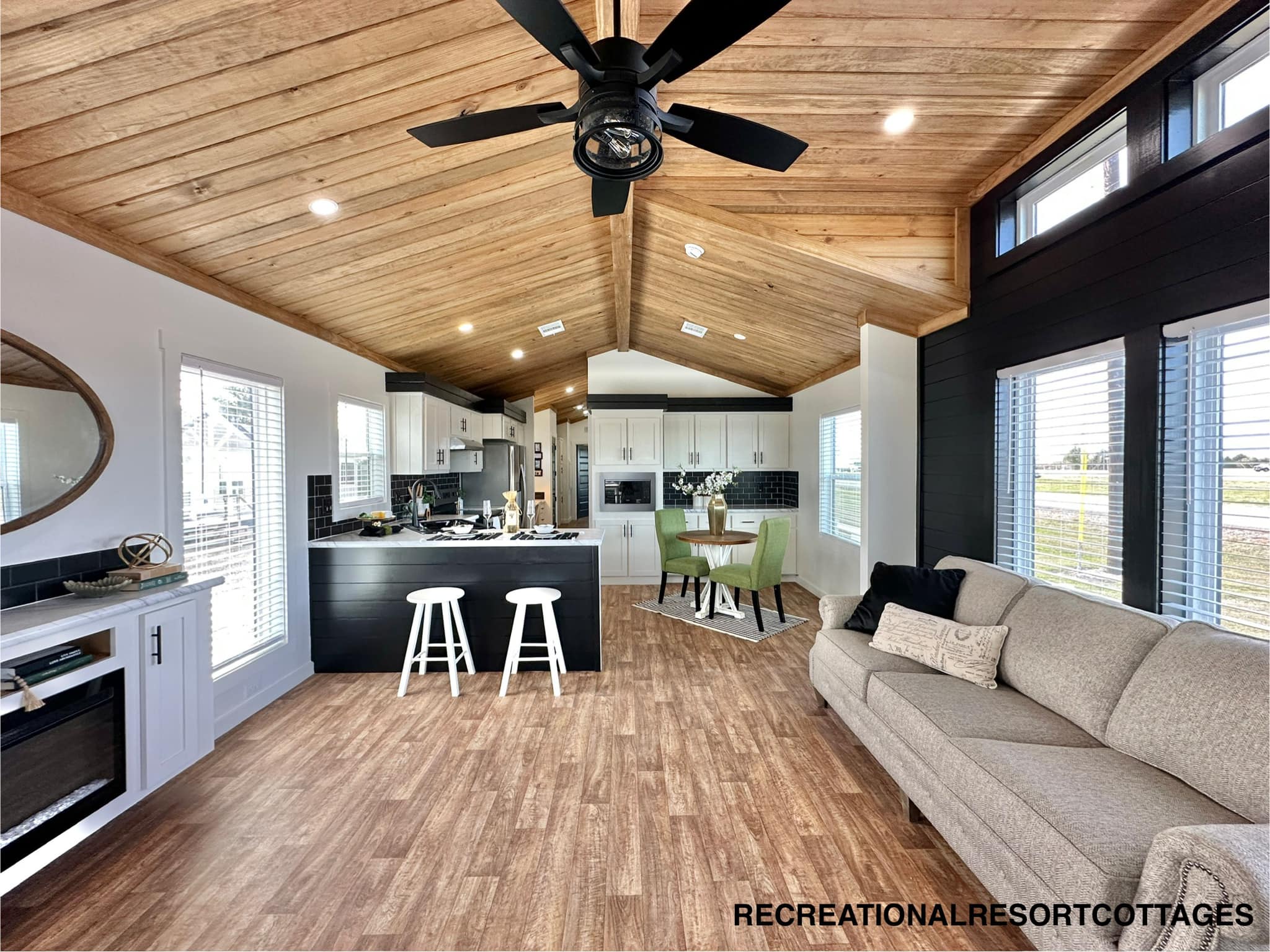 Stylish open-plan living area with wooden ceiling and floor, featuring a modern kitchen with black accents, cozy beige sofa, and a small dining set.