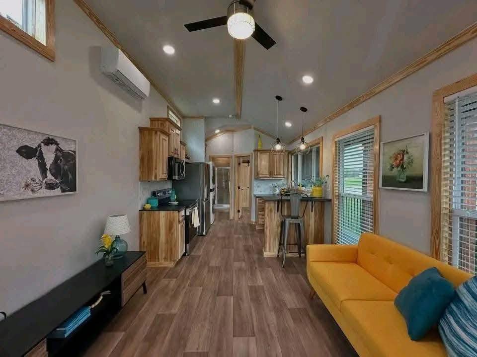 Modern open-plan living area featuring wooden floors, a bright yellow sofa, and a kitchen with wooden cabinets. Artwork and greenery add warmth.