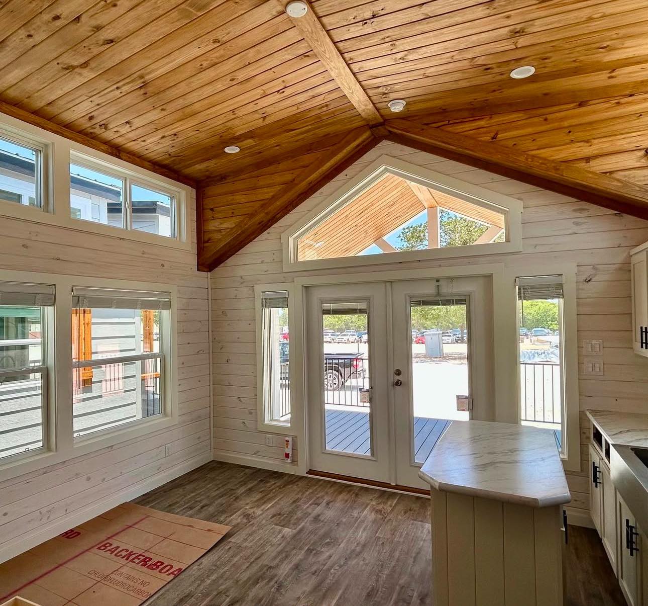 Cozy sunlit room with white wood-paneled walls and a pitched cedar ceiling. Large double doors and numerous windows lead to a bright outdoor view.