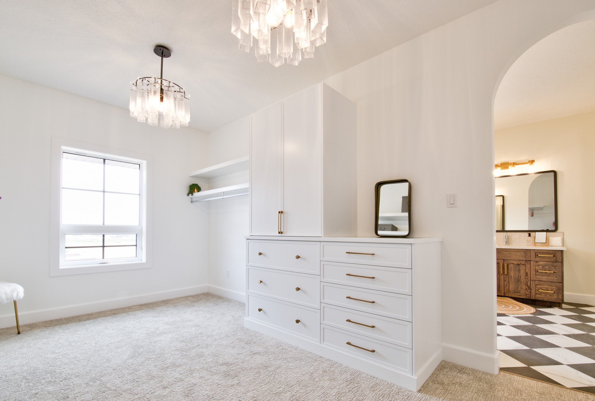 Spacious, modern walk-in closet with white cabinetry and gold handles. A small mirror rests on the dresser. An arched doorway leads to a bathroom.