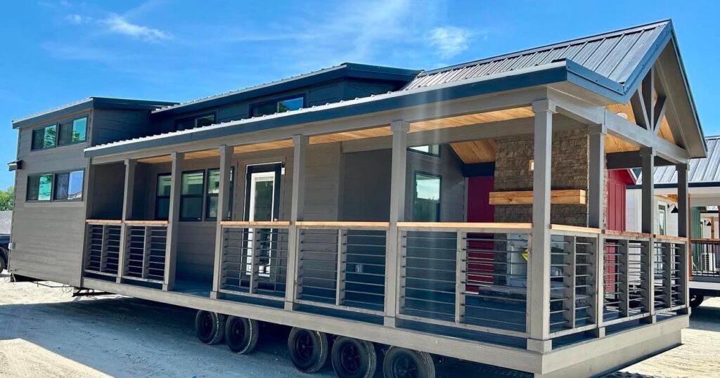 A modern gray tiny house on wheels with a covered porch, large windows, and a metal roof under a clear blue sky, evoking a sense of mobility and comfort.
