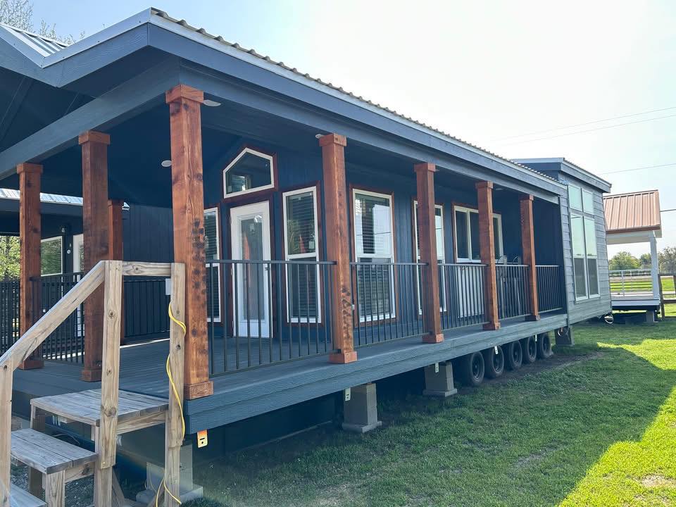 A modern, blue tiny house with large windows and wooden pillars stands on a grassy lawn. Stairs lead to a porch. The scene is bright and inviting.