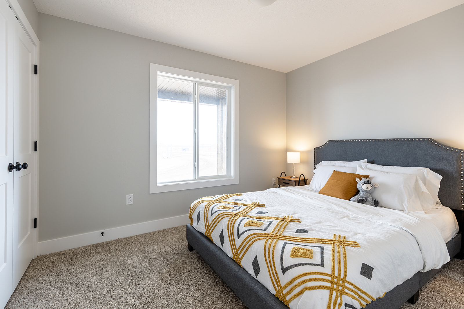 Cozy bedroom with a gray upholstered bed, white and mustard geometric bedding, and a stuffed raccoon. Soft lighting from a bedside lamp.