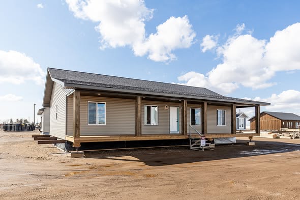 Single-story modular home with beige siding and a black roof, set against a bright blue sky with white clouds. The foreground shows a muddy construction site.
