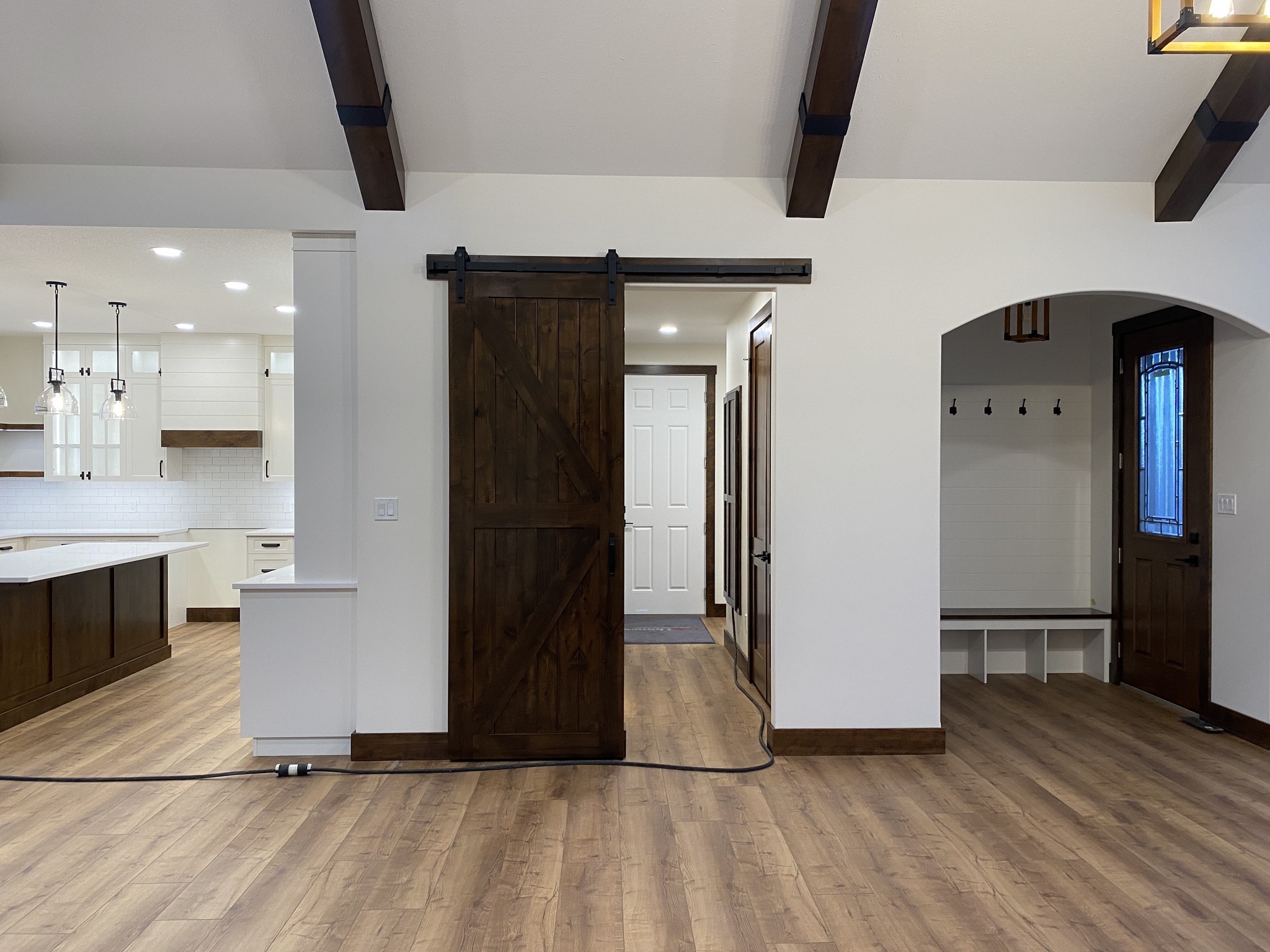 A cozy room features light wood flooring and dark wooden beams. A white kitchen with pendant lights is visible on the left. A central sliding barn door in dark wood adds rustic charm, leading to a hallway. An arched nook with coat hooks and a bench is on the right. The tone is warm and inviting.