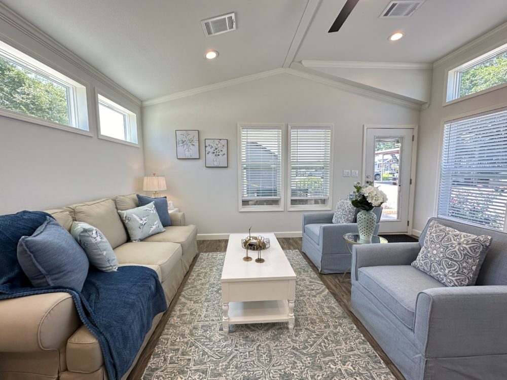 Bright living room with soft, neutral tones. Beige couch with blue cushions on the left, two blue armchairs on the right. White coffee table, floral decor, patterned rug, and wall art complete the cozy, inviting space.