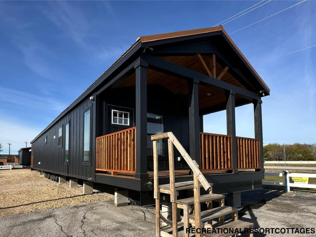 Modern tiny home with dark exterior and wood accents on porch, situated on gravel. Stairs lead to entrance, under clear blue sky, conveying simplicity.