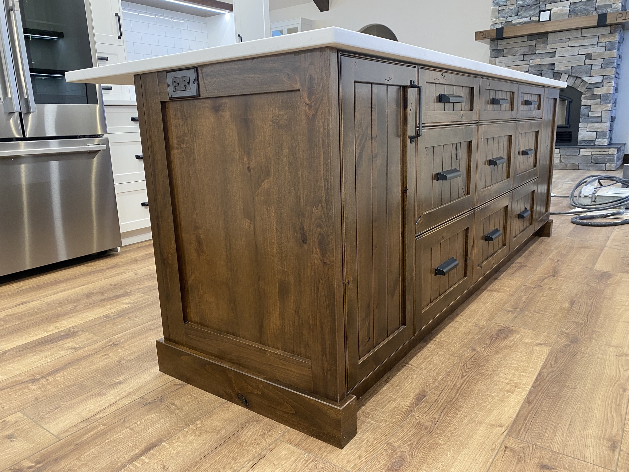 Wooden kitchen island with dark handles, set in a modern kitchen. Stainless steel appliances and stone fireplace in the background on a wood floor.