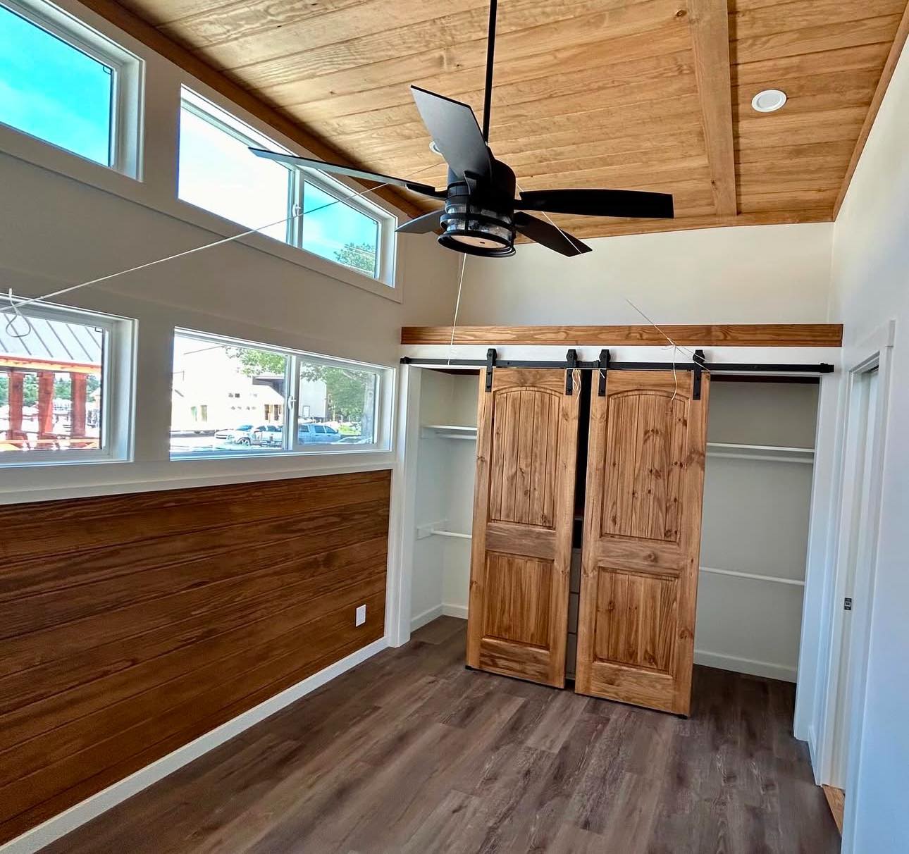 Bright room with wooden ceiling and flooring, featuring four narrow upper windows. A ceiling fan hangs above sliding barn doors on a closet, creating a cozy, modern feel.