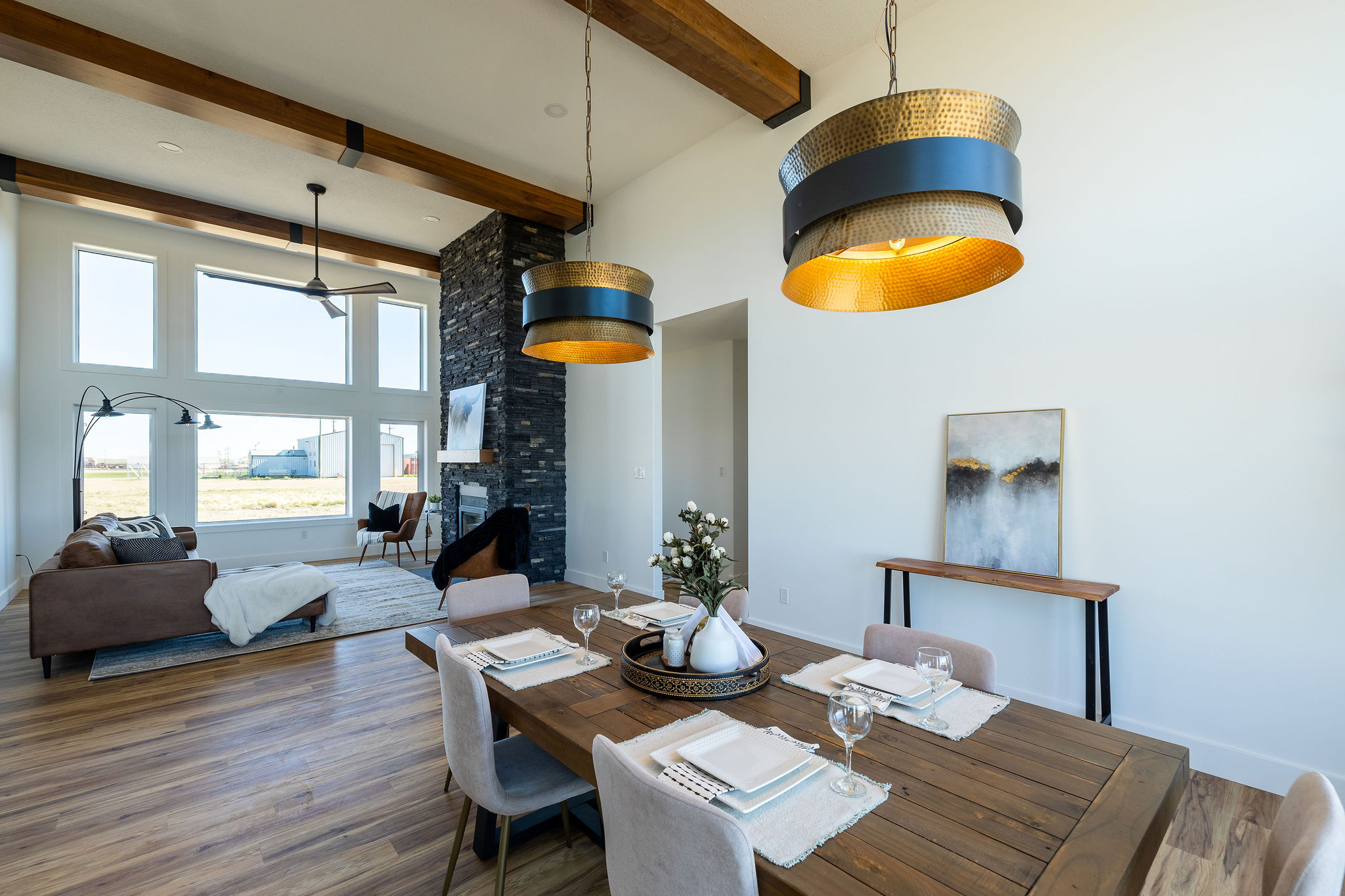 Spacious modern living area with high ceilings, wooden beams, and large windows. Features a cozy dining table, pendant lights, and stone fireplace.