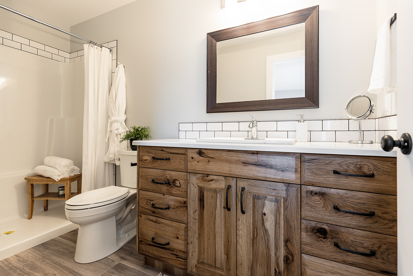 Cozy bathroom with wood vanity, large mirror, and white tiles. A toilet and shower with curtain sit beside a small wooden stool with towels, conveying warmth.