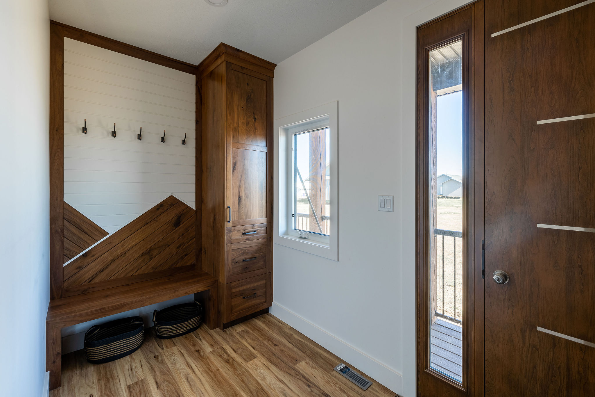 A modern mudroom with a wood bench and cabinet, four wall hooks, two storage baskets, a small window, and a wooden door. It feels warm and organized.