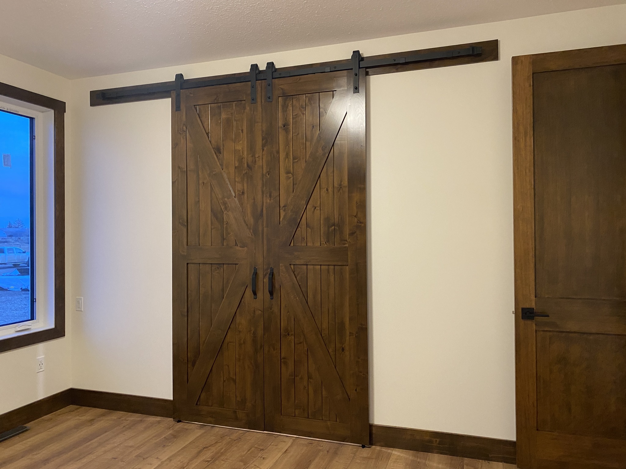 Wooden barn-style sliding doors with a dark finish are mounted on a black metal track in a cozy room with beige walls and wood flooring.