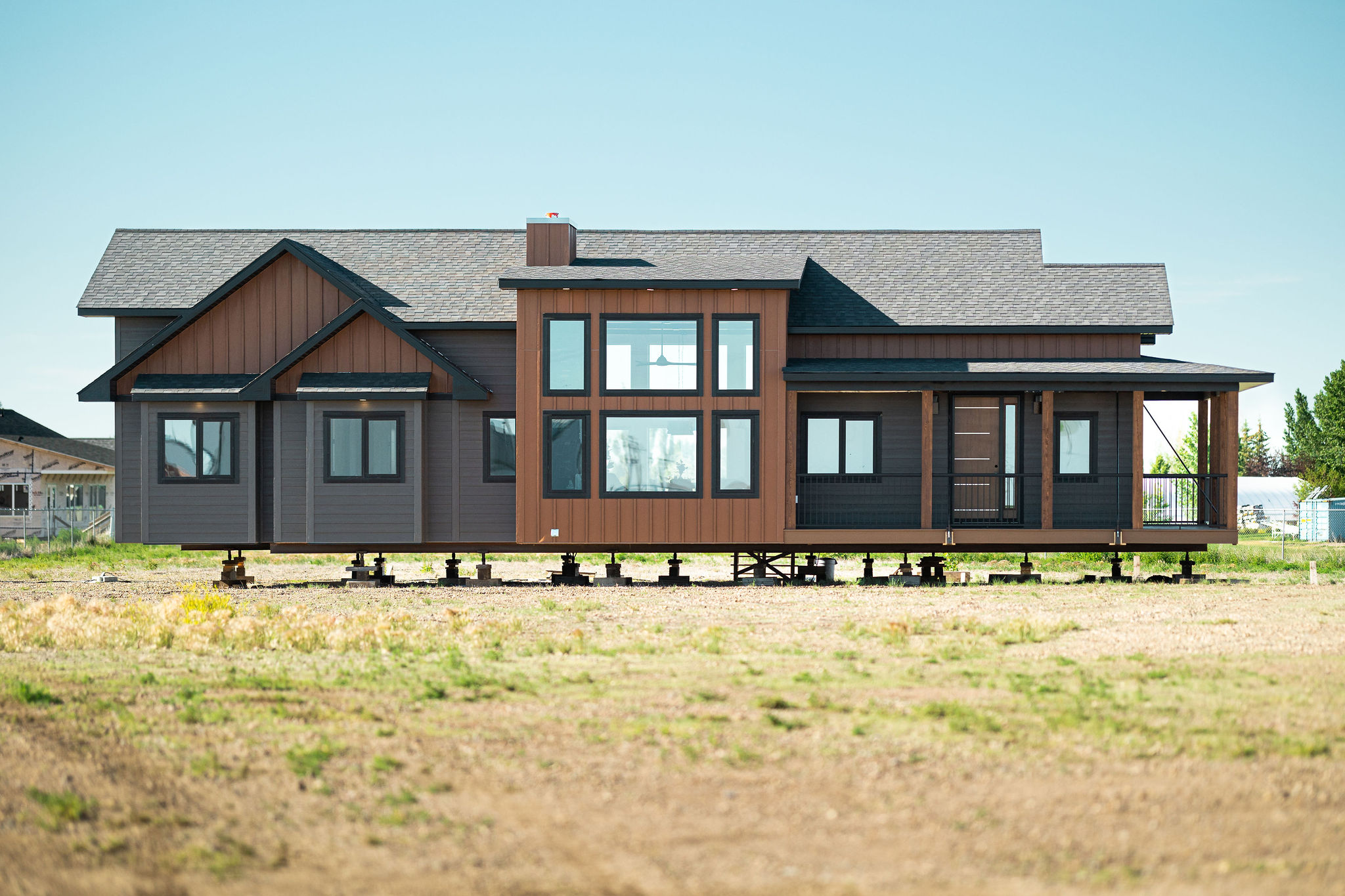 Modern prefab home elevated on a grassy plot, featuring large windows and a porch. Background includes a clear blue sky and distant trees.