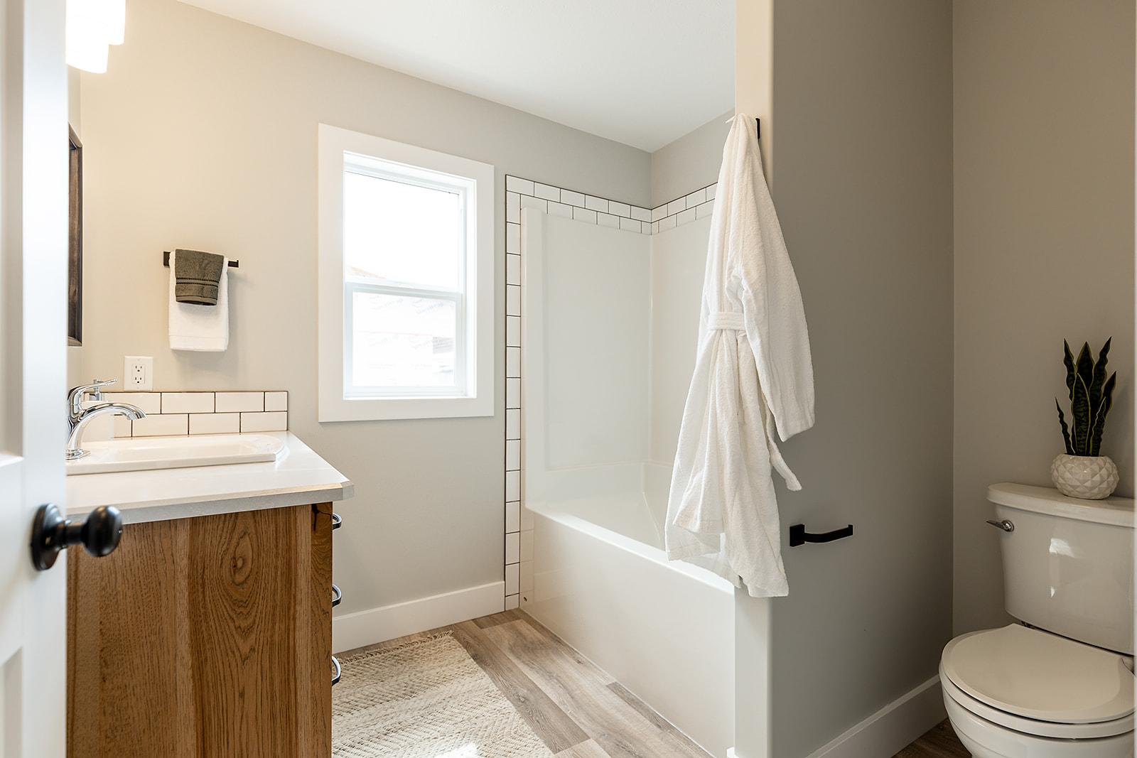 Bright bathroom with white bathtub and robe hanging on wall. Wooden vanity with sink, window, and light beige walls create a clean, inviting atmosphere.