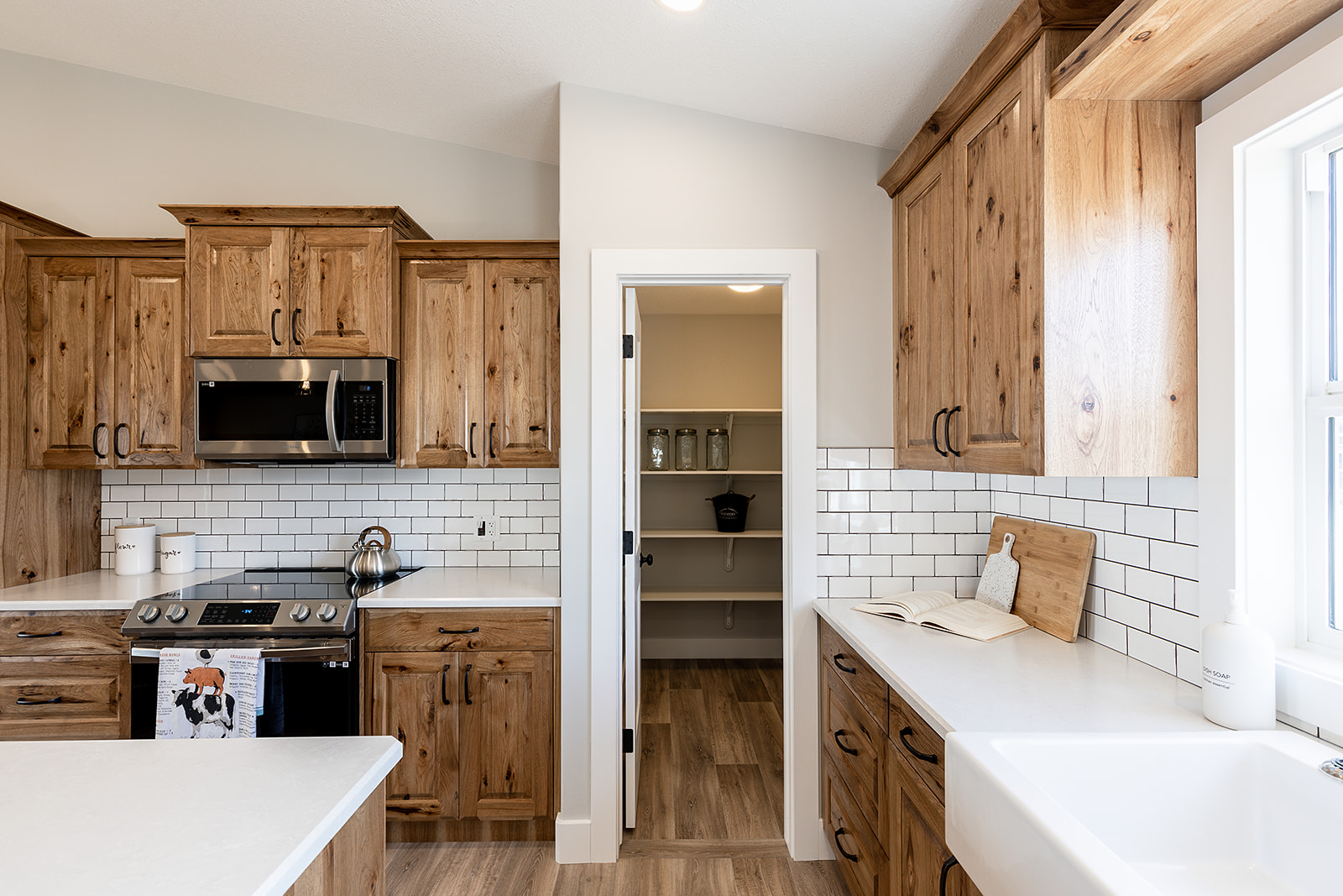 A modern kitchen with wooden cabinets, white subway tile backsplash, and stainless steel appliances. An open pantry is visible through a door. Bright and inviting.