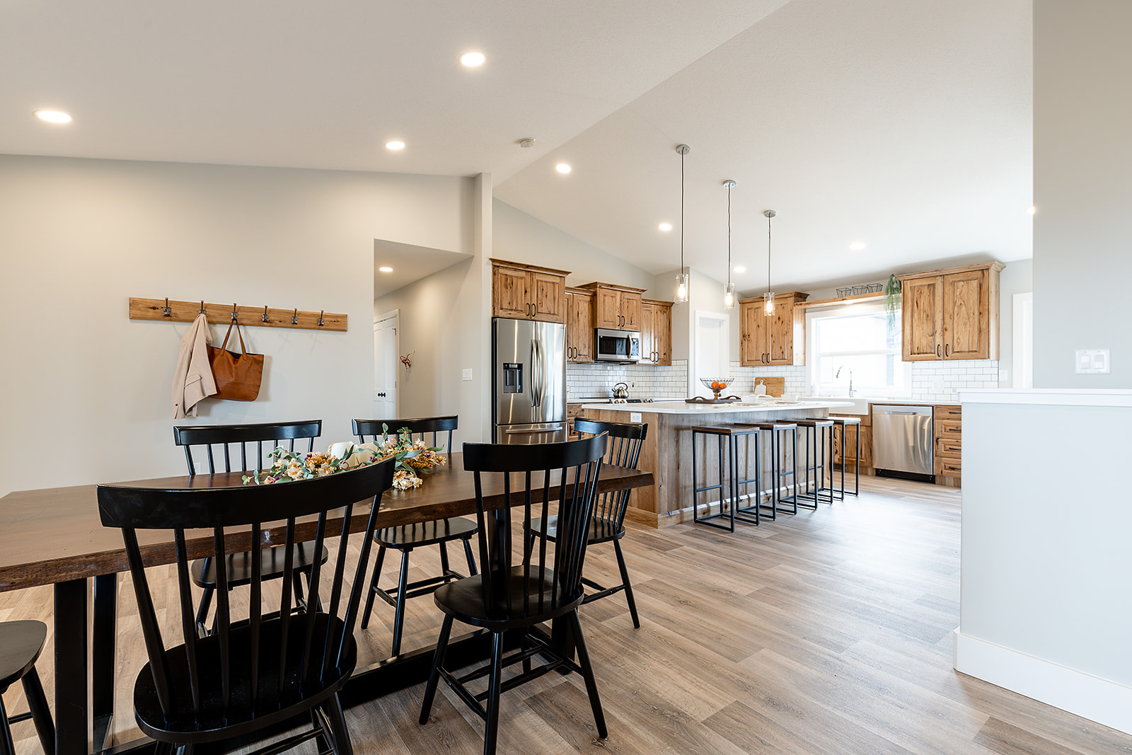Spacious kitchen and dining area with light wood floors, black chairs around a table, wooden cabinets, and a modern island with metal stools and pendant lights.