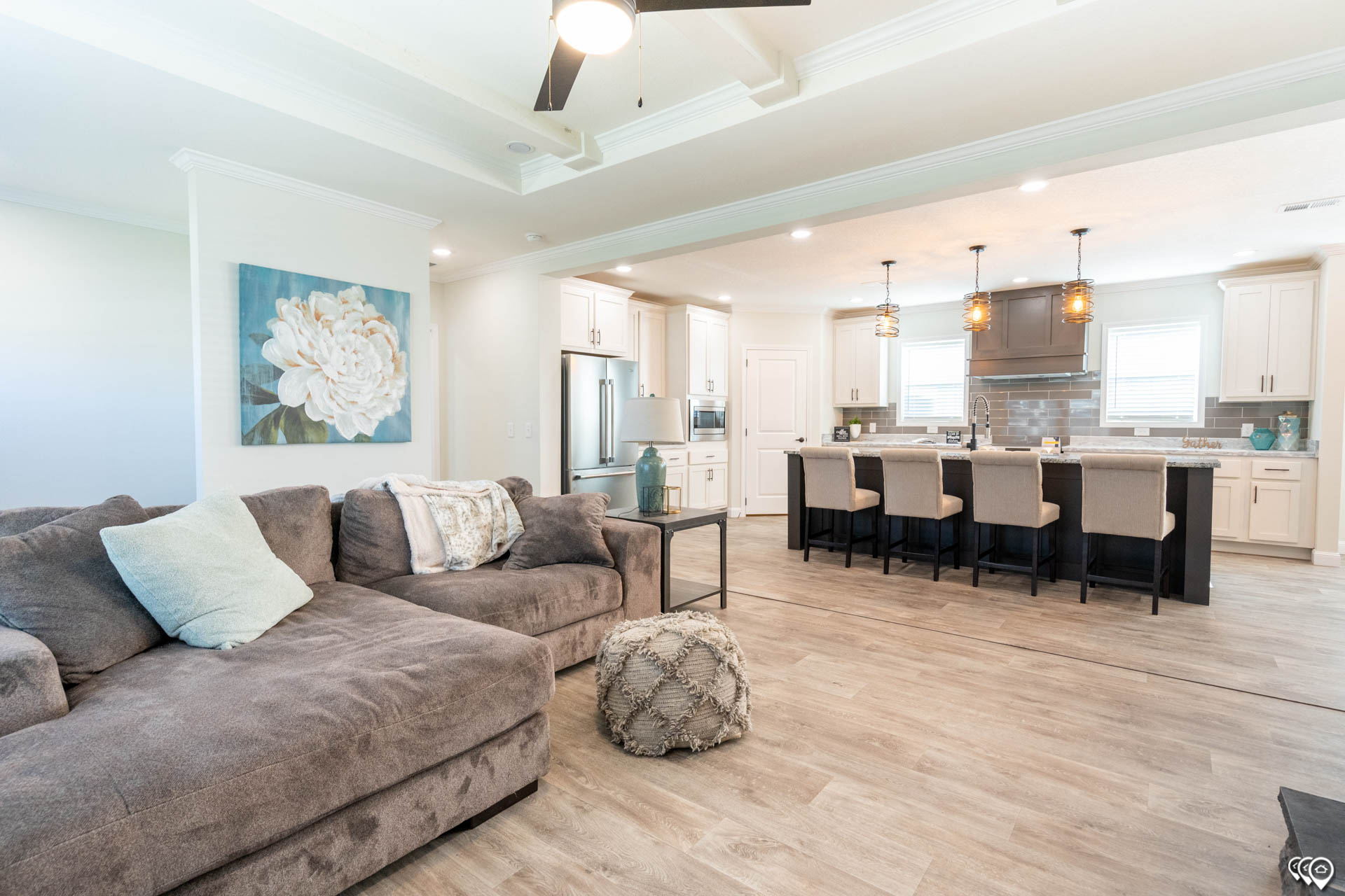 Spacious open-concept living room and kitchen with light wood flooring. Cozy gray sectional, accent pillows, a pouf, and elegant kitchen island with stools.