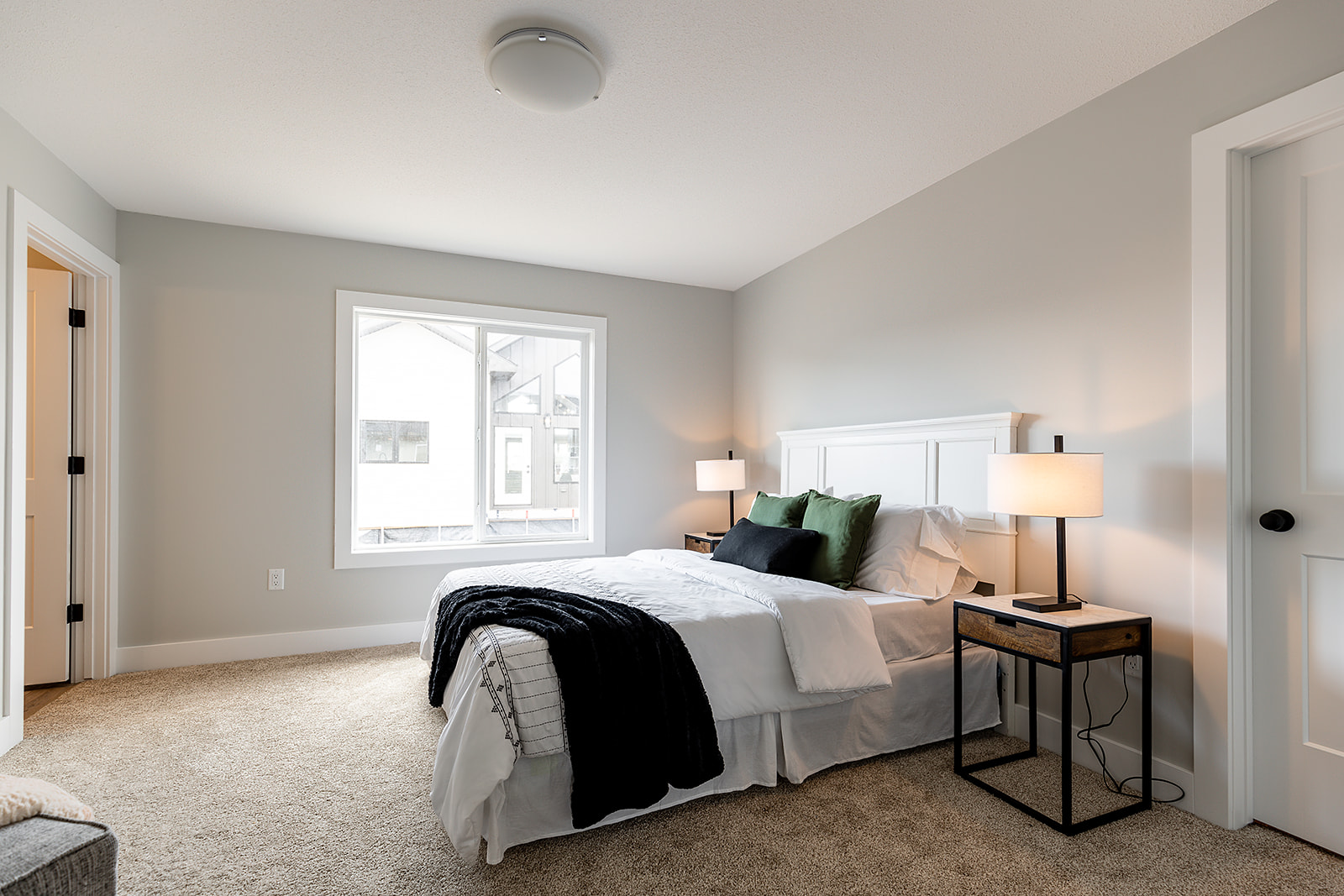Bright bedroom with a large window, gray walls, and beige carpet. A neatly made bed with white linens, green and black pillows, and bedside lamps create a serene and cozy atmosphere.
