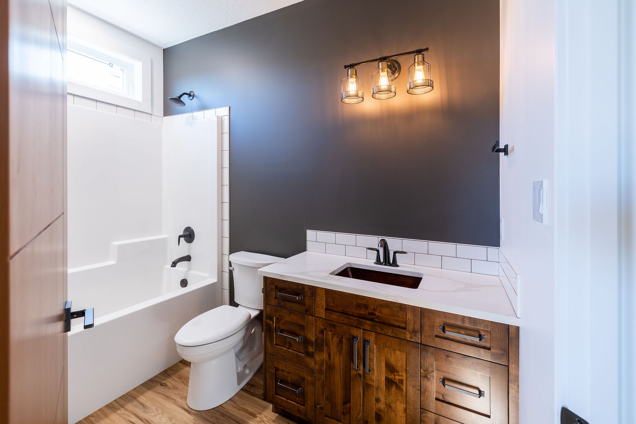 Bright, modern bathroom with rustic wood vanity, white tub, and toilet. Gray accent wall with vintage light fixtures adds warmth. Simple and clean design.