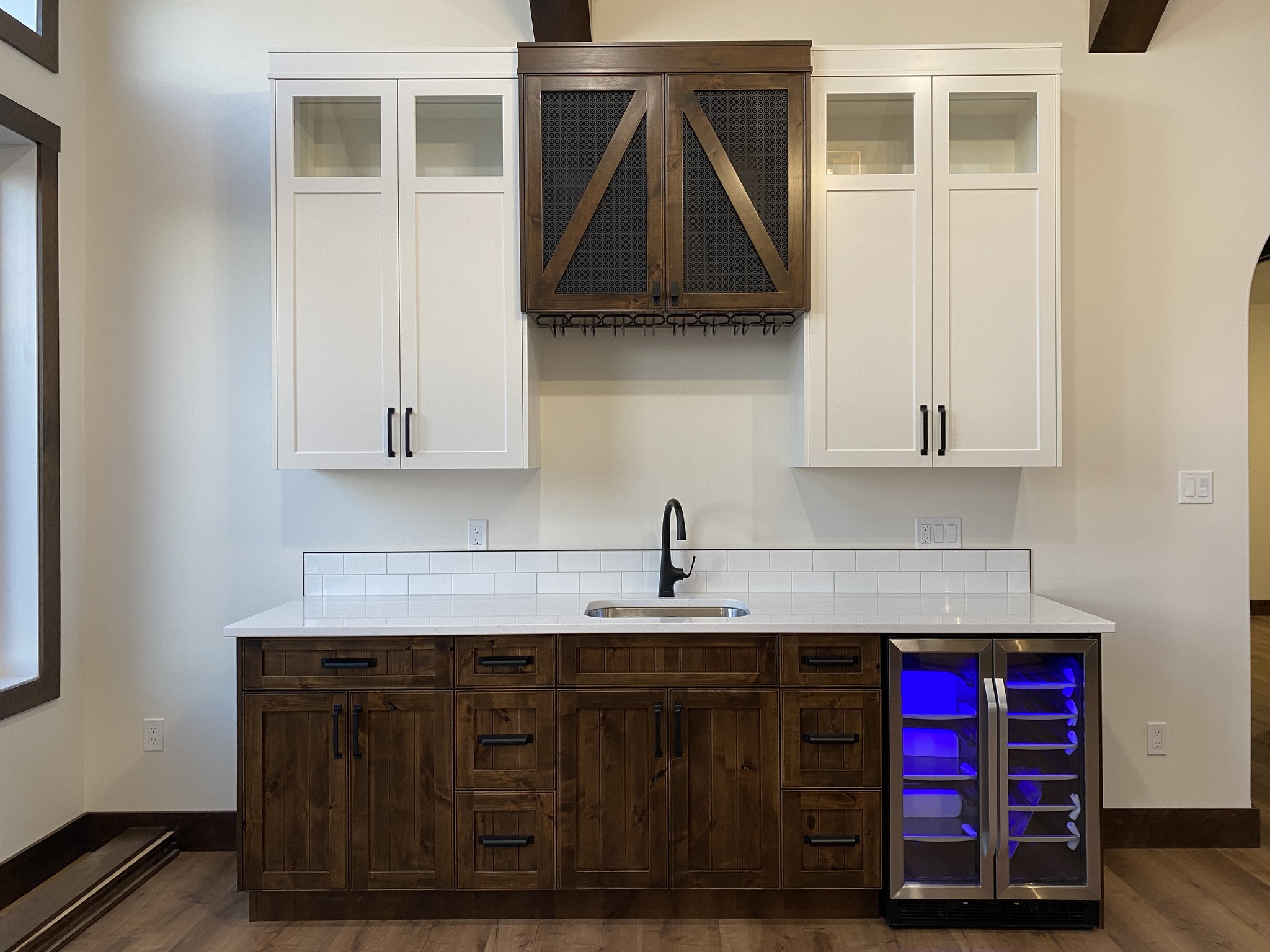 Modern kitchen with white cabinets, wooden accents, black handles, and a sleek black faucet. A wine fridge with blue lighting completes the stylish look.