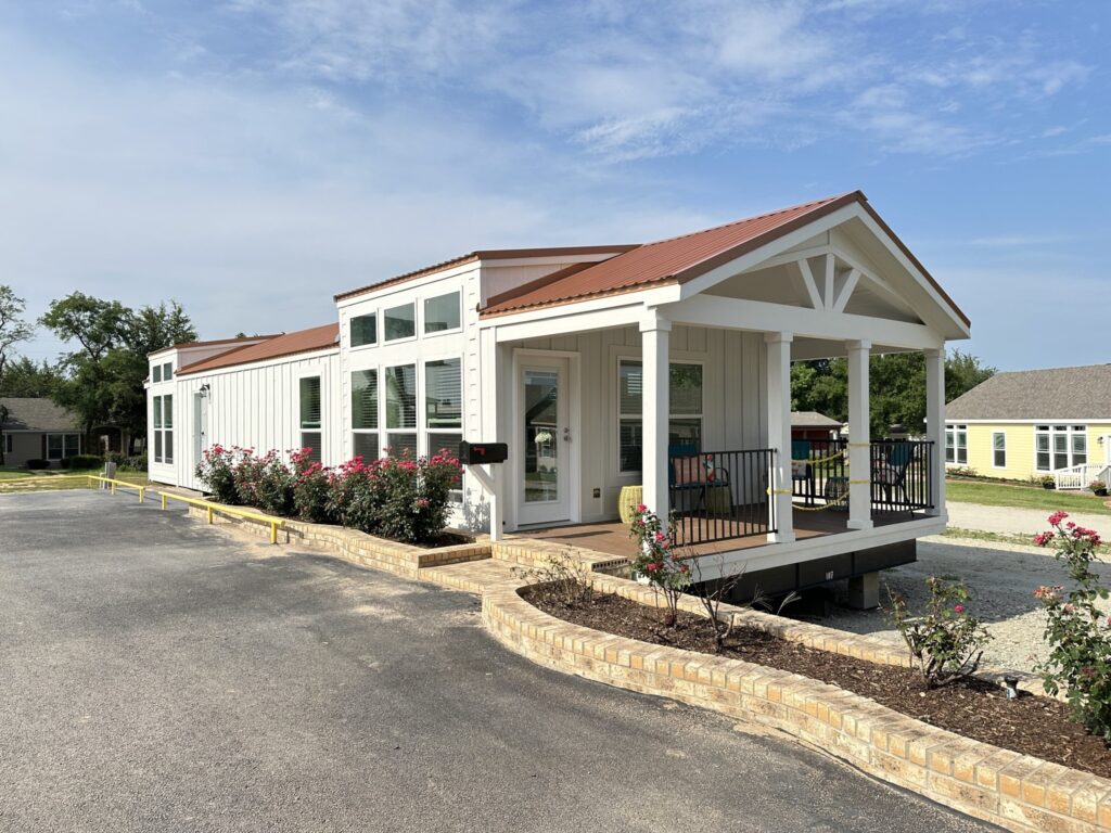 A small, modern white house with a red roof and large windows. The front porch is adorned with flowers, exuding a cozy, inviting ambiance under a bright sky.