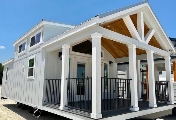 A modern tiny house with white siding, a covered porch, black railings, and large windows against a clear blue sky, conveying a cozy and inviting atmosphere.