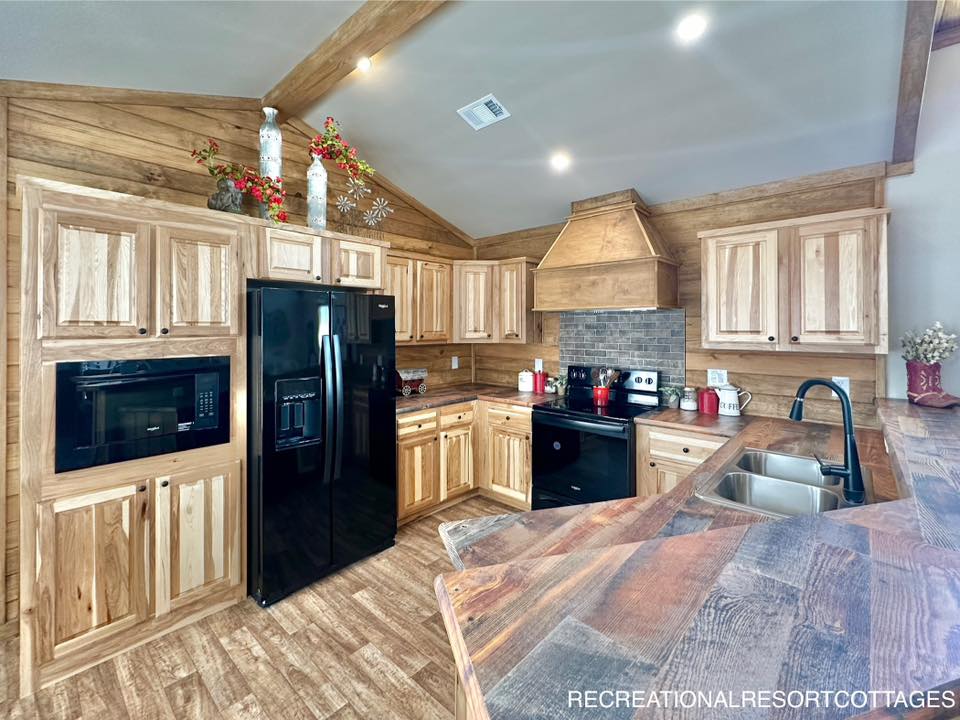 Rustic kitchen with wood cabinets, black appliances, and a brick backsplash. Decorative vases with red flowers adorn the wood-paneled walls. Warm and cozy ambiance.