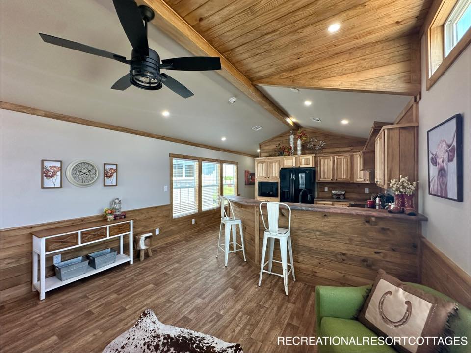 Spacious rustic living room with wood accents, ceiling fan, and natural light. Features a kitchen with bar stools, cow-themed decor, and cozy seating.