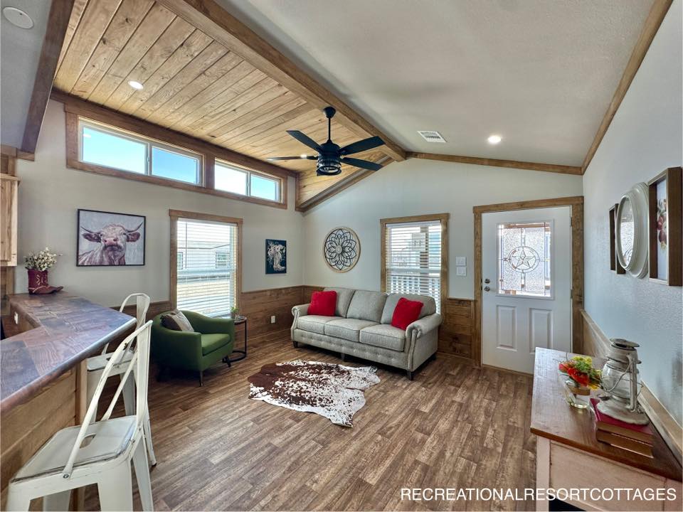 Cozy living room with wood accents, featuring a beige sofa with red pillows, a green armchair, and a cowhide rug. Bright natural light fills the space.