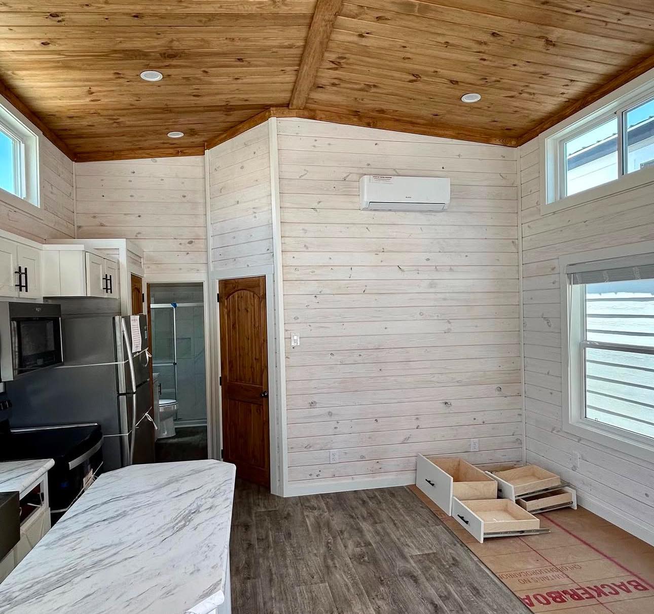 Cozy room with wood-paneled walls and ceiling, featuring a modern kitchen with stainless steel appliances, marble countertops, and natural light streaming through windows. A wooden door and storage drawers create a warm, welcoming atmosphere.