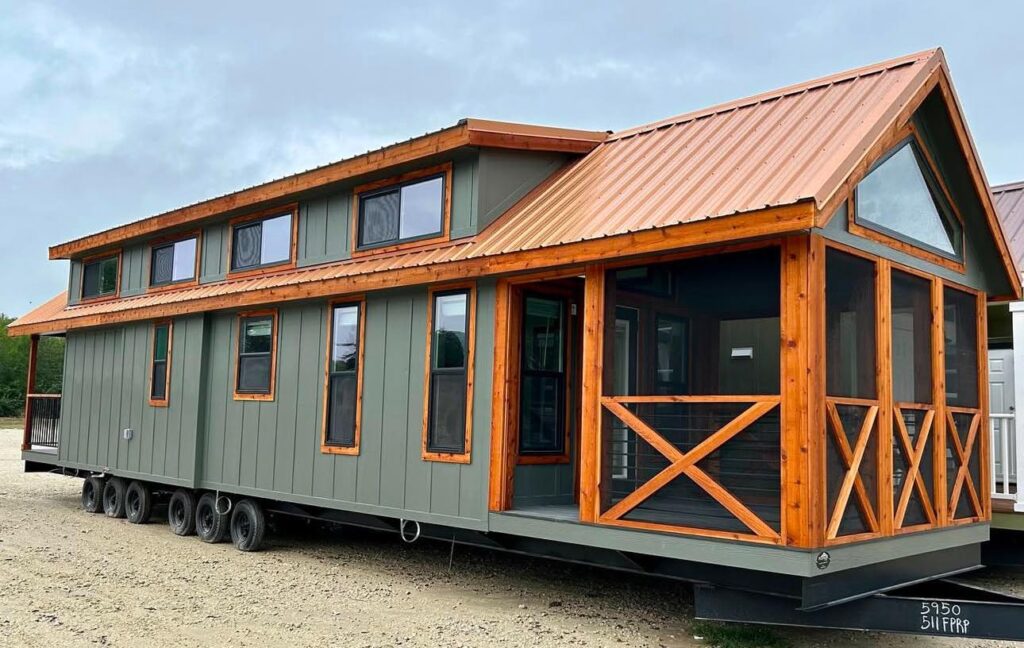 A modern tiny house on wheels with green siding and orange wooden trim. It features a gabled roof, large windows, and a porch with cross-braced railings.