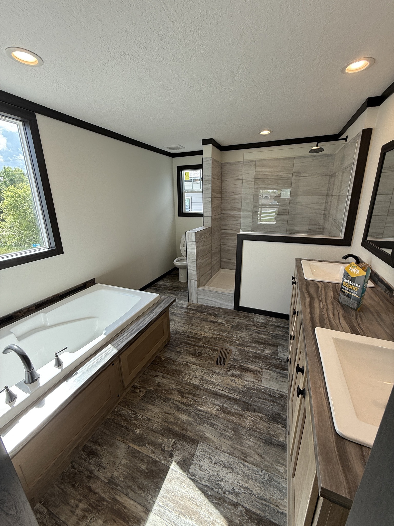 Modern bathroom with wood-tone flooring, a large white bathtub on the left, walk-in shower with gray tiles, and dual sinks on a dark countertop.