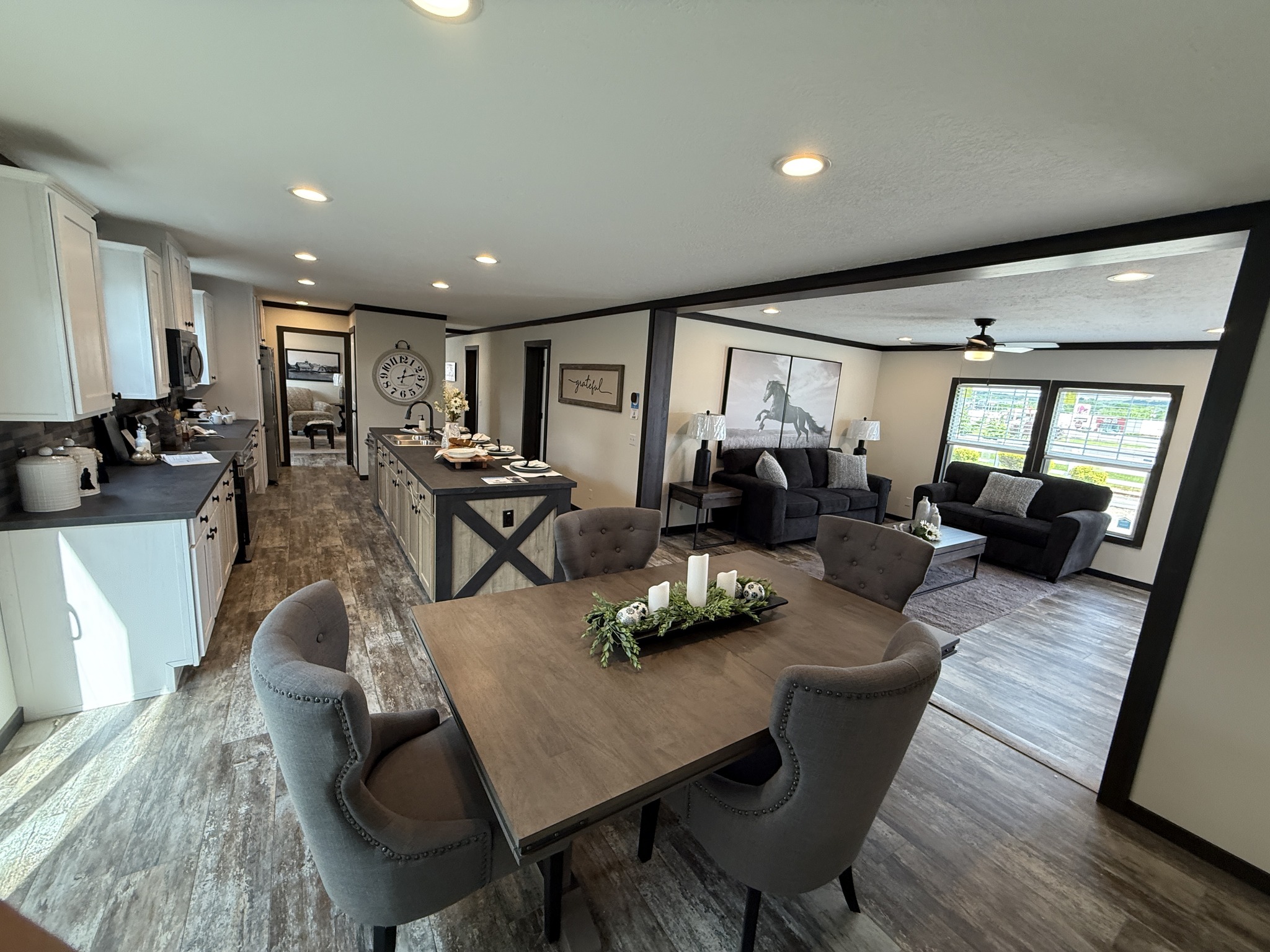Modern open-plan kitchen and living area with rustic decor. Features a dining table with candles, gray sofas, and a wall-mounted horse artwork. Bright and cozy ambiance.