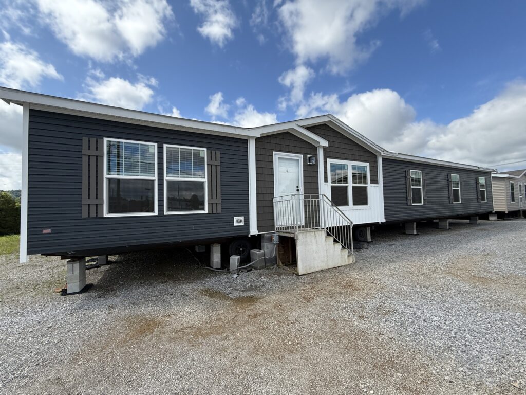 Gray manufactured home with white trim, elevated on concrete blocks. It has several windows and a front door with steps, under a partly cloudy sky.