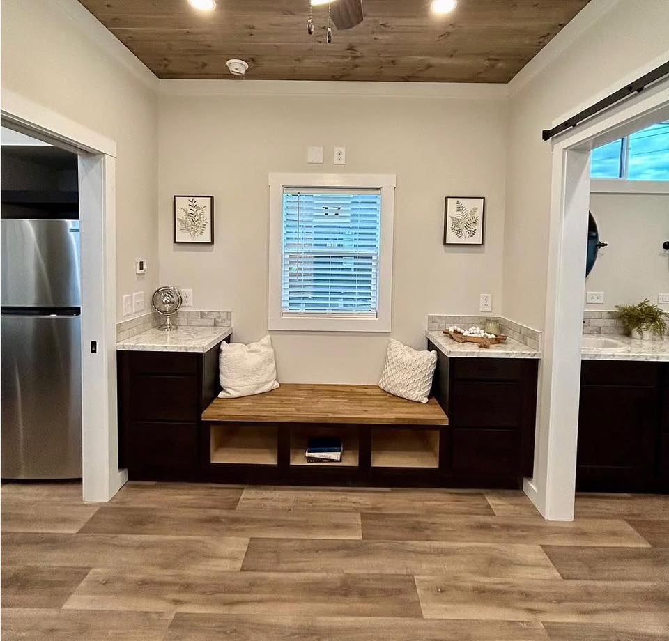 Bright room with wooden floors and ceiling, featuring a cozy bench with white pillows under a window. Marble-topped dark cabinets flank the bench.
