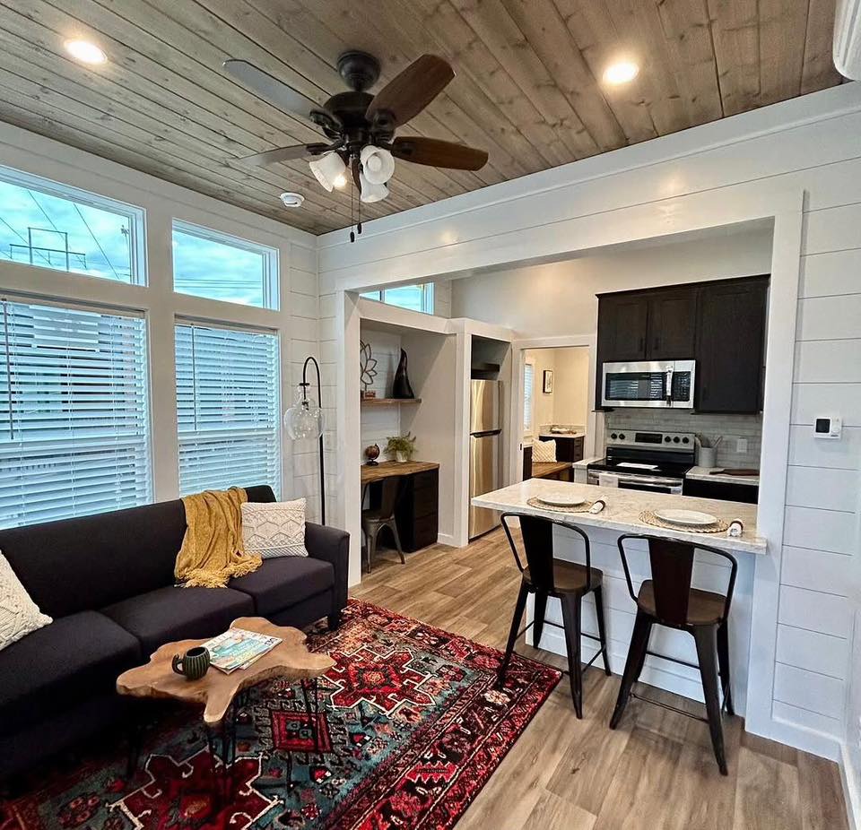 Cozy living area with a dark sofa, colorful red rug, and natural wood coffee table. Open kitchen features bar stools and modern appliances. Warm, inviting ambiance.