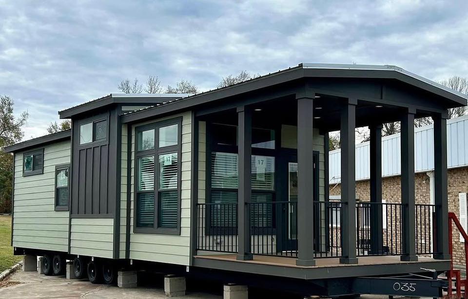 Modern tiny home on wheels with light green siding and dark trim. Features large windows, a small front porch with railing, set in a park-like setting.
