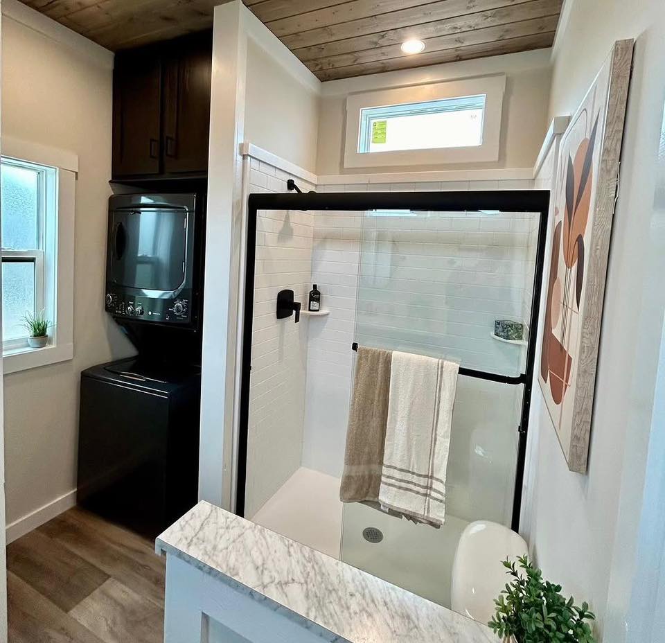 Modern bathroom with a wood ceiling, featuring a glass shower with a towel hanging, a stacked washer and dryer, and warm wooden flooring.