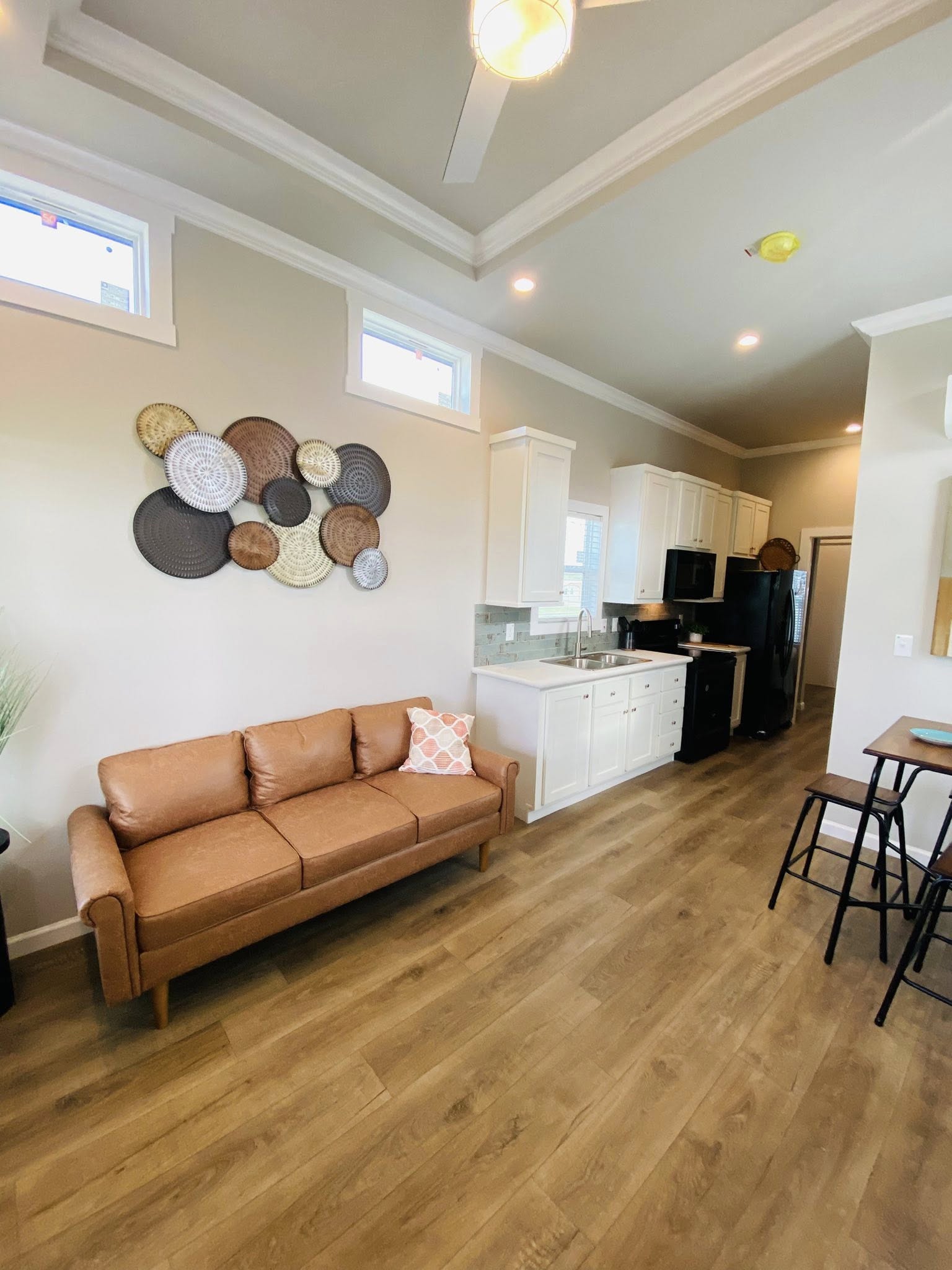 Modern living room with a brown leather sofa, decorative woven wall art, and a sleek kitchen featuring white cabinets and stainless steel appliances. Cozy and inviting.
