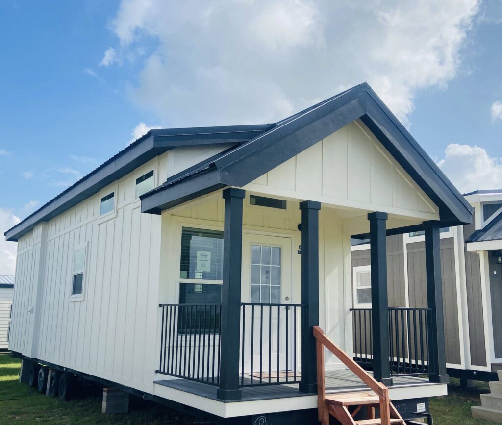 A tiny house on wheels with a white exterior and dark trim sits under a partly cloudy sky. It features a small porch with railing and wooden A tiny house on wheels with a white exterior and dark trim sits under a partly cloudy sky. It features a small porch with railing and wooden steps.steps.