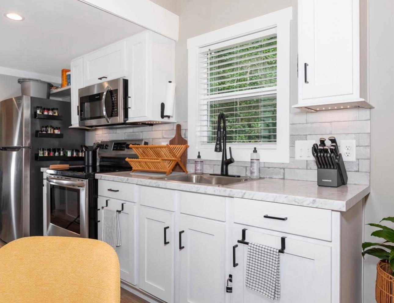 Bright kitchen with white cabinets, marble countertops, and stainless steel appliances. A wooden dish rack and knife set rest beside the sink. Airy and modern.