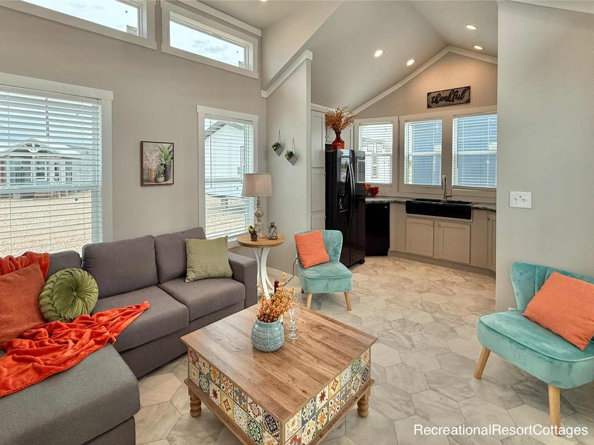 Cozy bright modern farmhouse open-concept living room and kitchen in a recreational resort cottage featuring vaulted ceiling with exposed beams, high clerestory windows flooding natural light, neutral gray walls and hexagonal tile flooring, plush gray sectional sofa with vibrant orange throw blanket and green pillows, turquoise accent chair, colorful tiled coffee table, wooden side table with lamp, black farmhouse sink, beige shaker cabinets, stainless steel refrigerator, "thankful" wall sign, potted plants and dried floral arrangements creating a warm inviting family space perfect for tiny home living, vacation cottages, or relaxed getaway retreats.