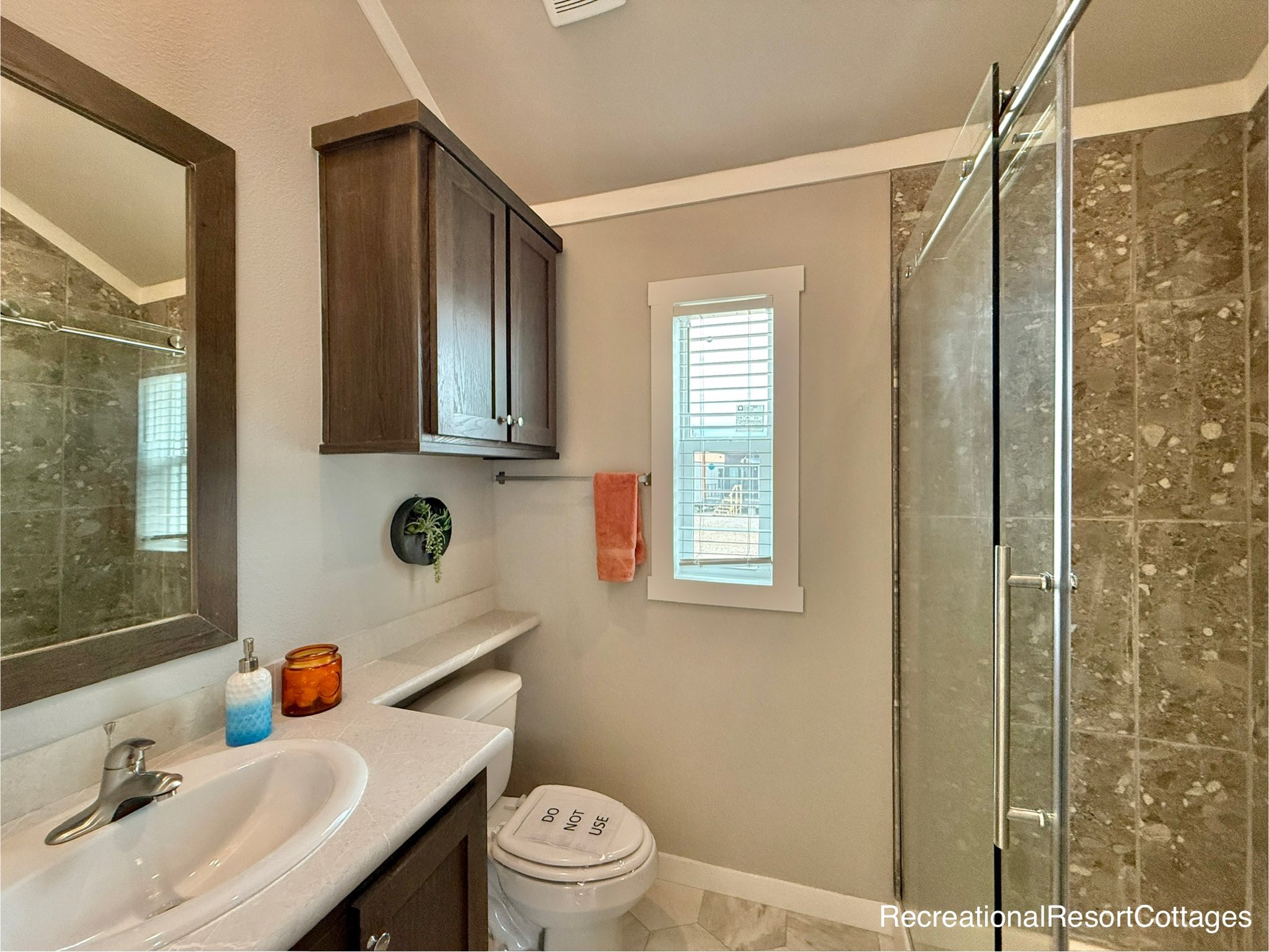Cozy modern farmhouse bathroom in recreational resort cottage featuring warm beige walls with textured stone-look tile surround, dark walnut vanity cabinet with white quartz countertop and undermount sink, large framed mirror, wall-mounted dark wood upper cabinet, walk-in glass shower with chrome hardware and neutral pebble tile flooring, hexagonal floor tiles, small high clerestory window for natural light, orange towel on chrome rack, potted succulent decor, amber glass jar accents, blue ombre soap dispenser, vaulted ceiling with recessed lighting, compact serene spa-like design perfect for tiny home living, vacation cottage retreats, or relaxing family getaways.