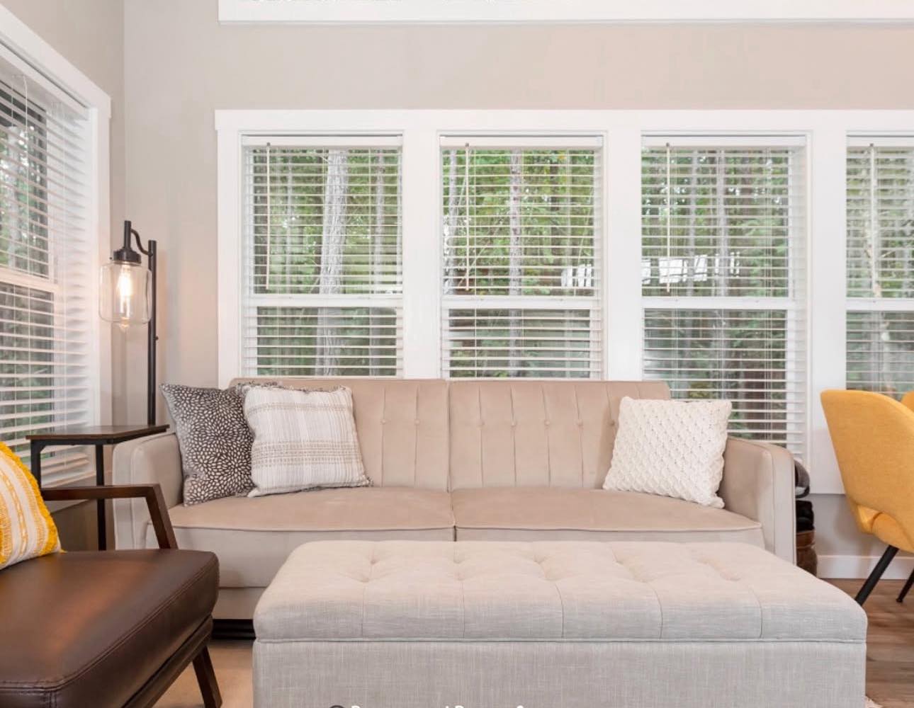 Stylish living room with a beige sofa and pillows in front of large windows with blinds. An upholstered ottoman and yellow chair add warmth.