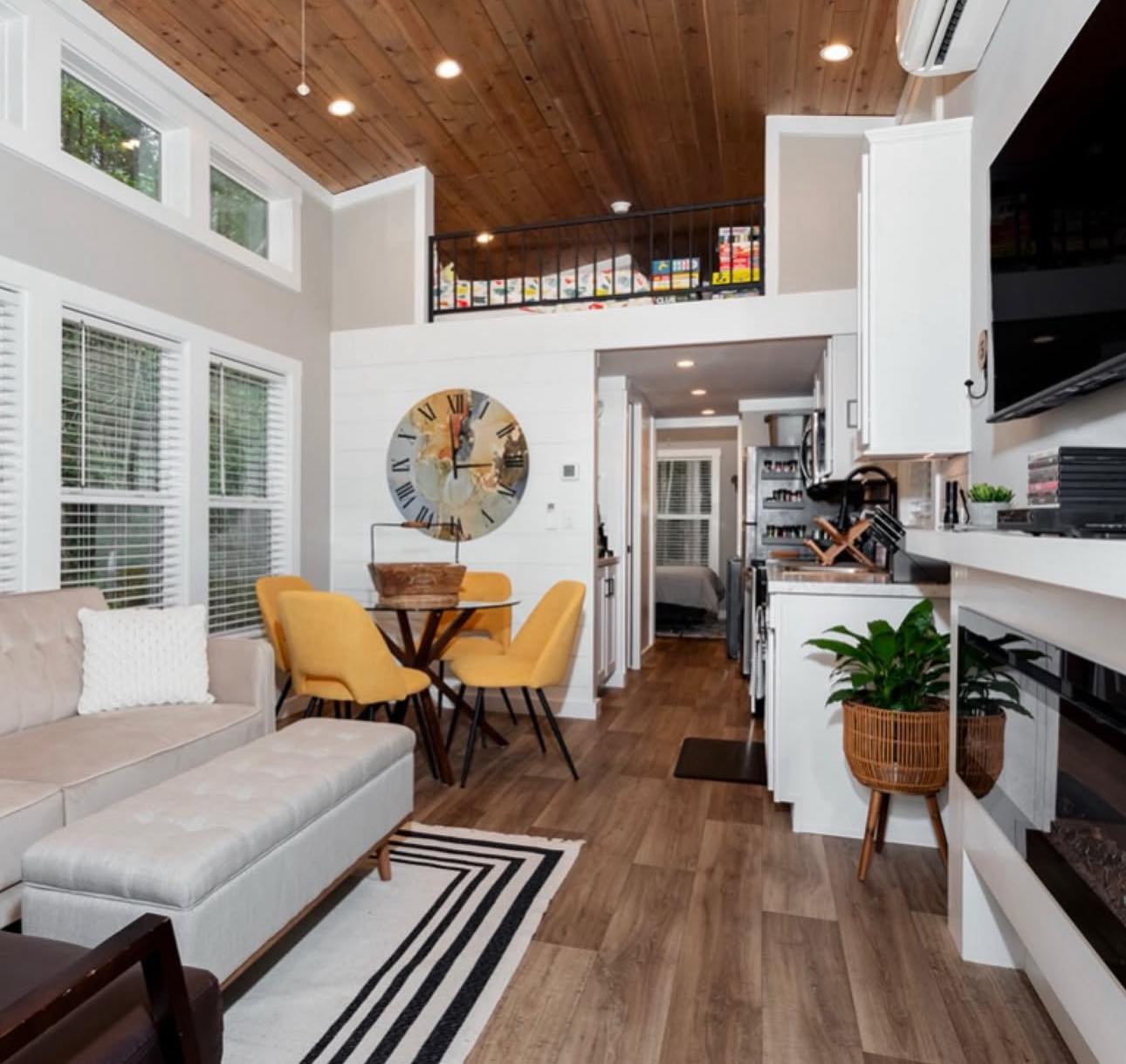 Cozy modern tiny home interior with wood ceiling, beige couch, yellow chairs around a round table, clock on wall, sleek kitchen, and lush plant decor.