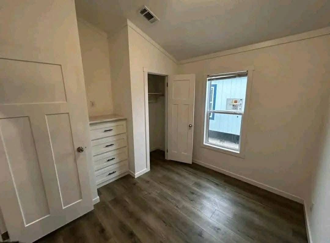 A small, bright room with light wooden flooring and white walls. It features a closet with drawers and a window letting in natural light. The atmosphere is clean and minimalist.