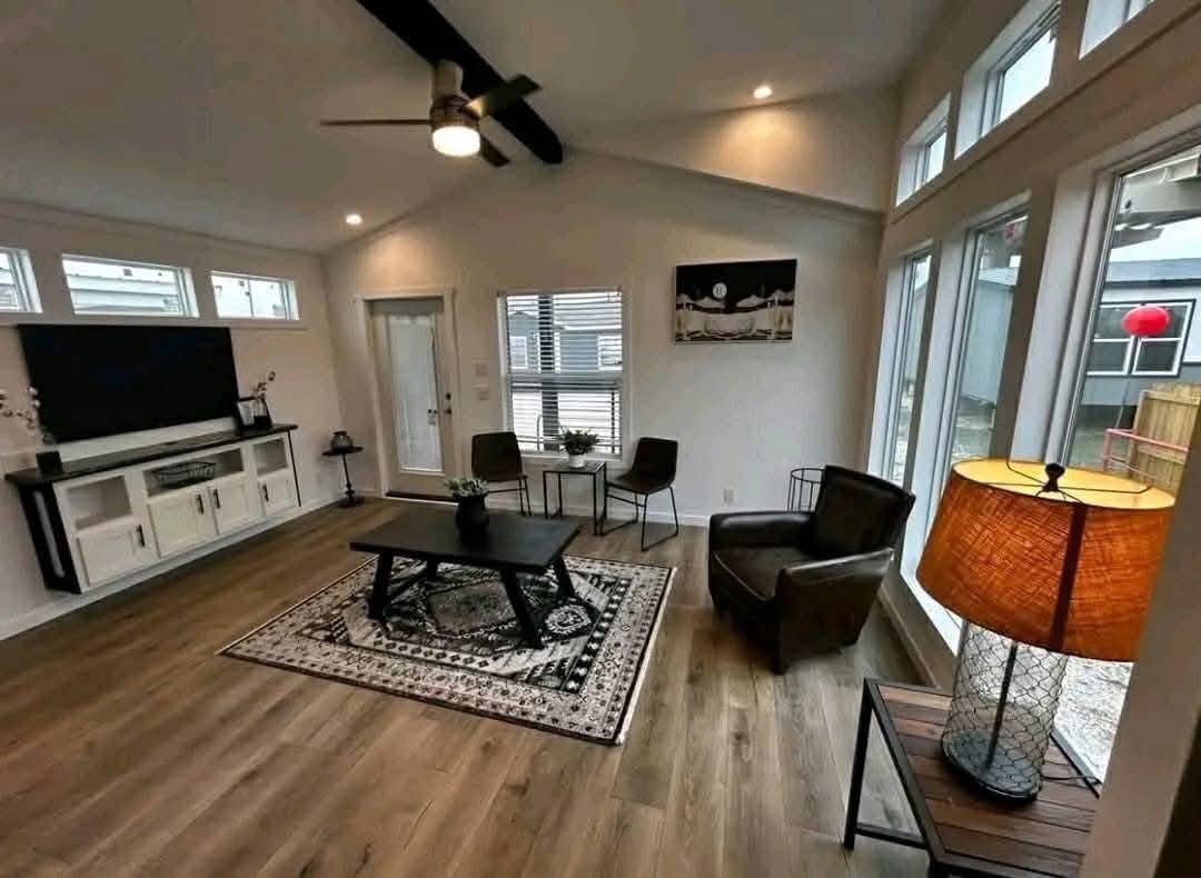 Modern living room with large windows, wood flooring, a ceiling fan, dark furniture, and a large patterned rug. Cozy, inviting atmosphere.