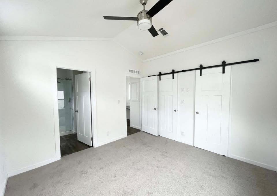 Bright, empty room featuring white walls, a carpeted floor, a ceiling fan, and sliding double doors. An open doorway leads to a tiled bathroom. Calm ambiance.
