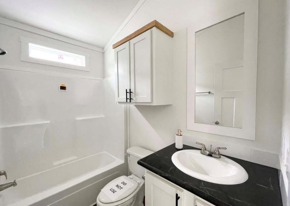 Compact, white bathroom featuring a bathtub, wall cabinet, and black counter with sink. A note on the toilet reads "Do Not Use," adding a cautionary tone.